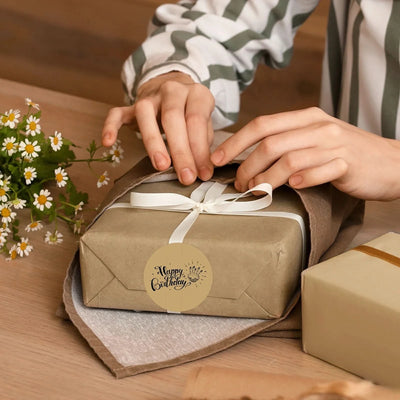 Person wrapping a birthday gift with a Happy Birthday label and white ribbon on a table