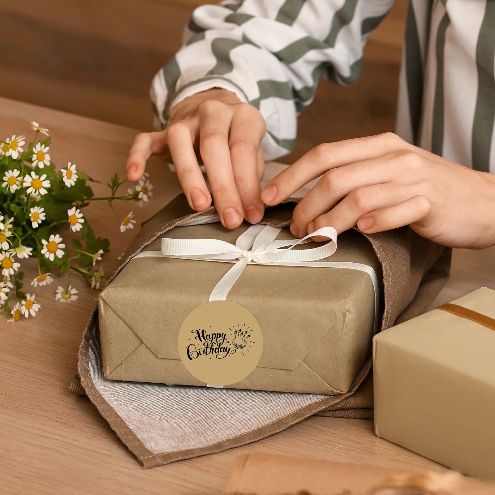 Person wrapping a birthday gift with a Happy Birthday label and white ribbon on a table