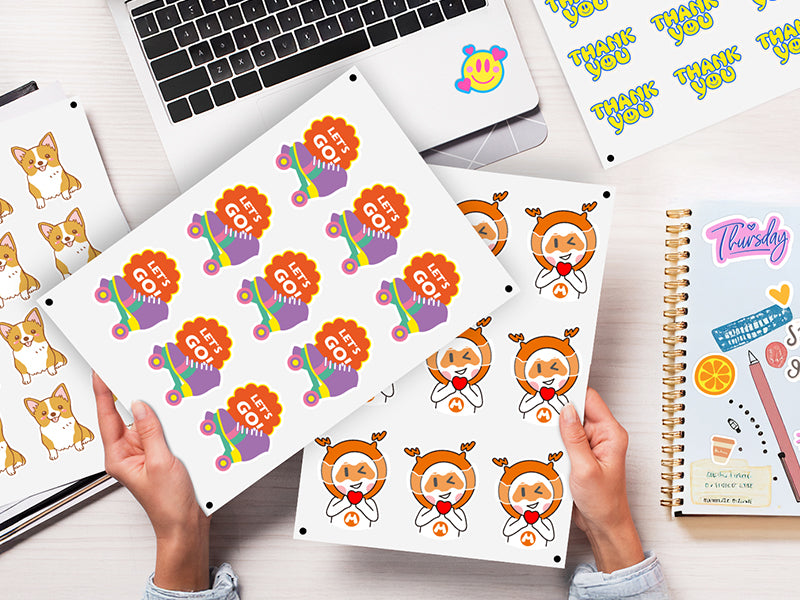 Sticker sheets with cartoon designs held by hands on a desk with a laptop and notebook.